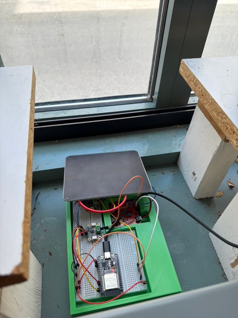 Plug-In Solar prototype positioned beside a window with its solar panel, green 3D-printed base, ESP32, sensor boards, battery charger, and wiring visible